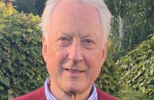 An older man stands on grass, wearing a red sweater and blue pants, with hedges and trees in the background.