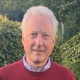 An older man stands on grass, wearing a red sweater and blue pants, with hedges and trees in the background.