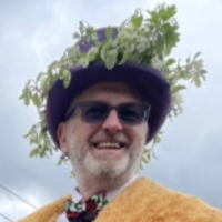 Smiling man wearing a purple hat decorated with green leaves and sunglasses, against a cloudy sky.