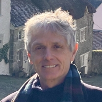 Smiling person with short gray hair and scarf standing outdoors in front of old stone cottages.