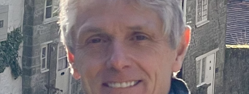 Smiling person with short gray hair and scarf standing outdoors in front of old stone cottages.